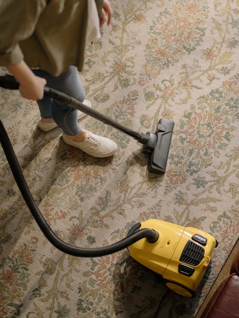 A person using a vacuum cleaner for surface cleaning on a patterned carpet inside a room. The individual is wearing beige shoes and jeans, operating a yellow and black vacuum cleaner with a long hose